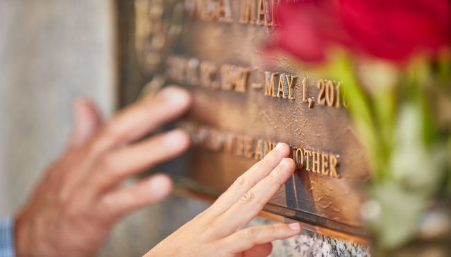 Hands reach out and touch a bronze marker at Rose Hills